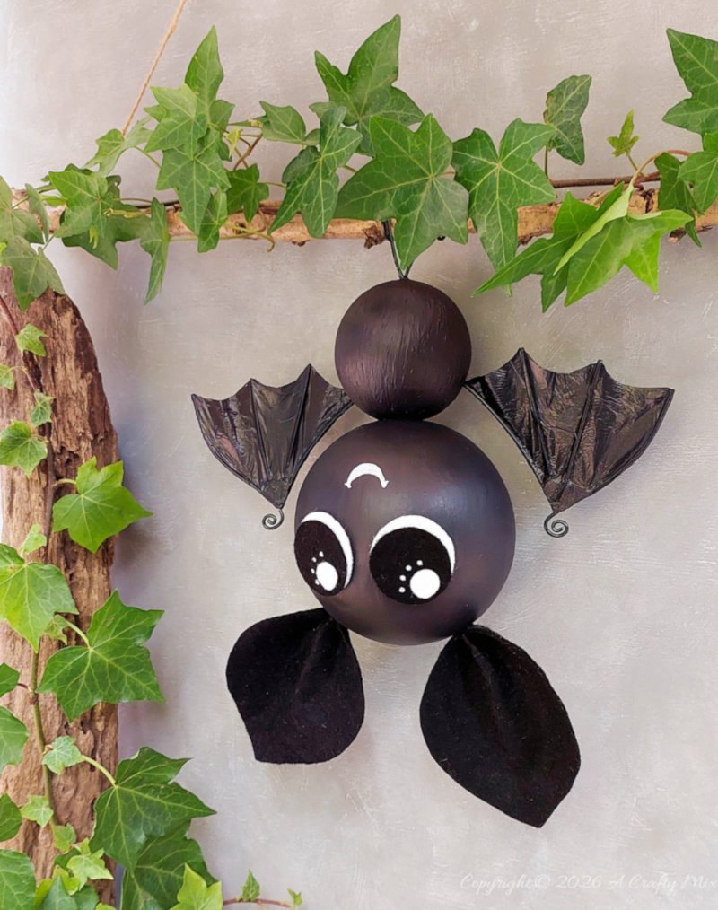 Cute black hanging Halloween bat with round eyes, shiny recycled plastic bag wings, and wire feet displayed beneath a leafy branch.