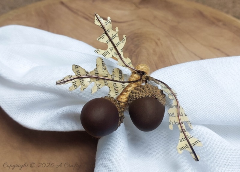 Close-up of handmade DIY acorn napkin rings with clay acorns, real acorn caps, copper wire, and book-page leaves on a white napkin.