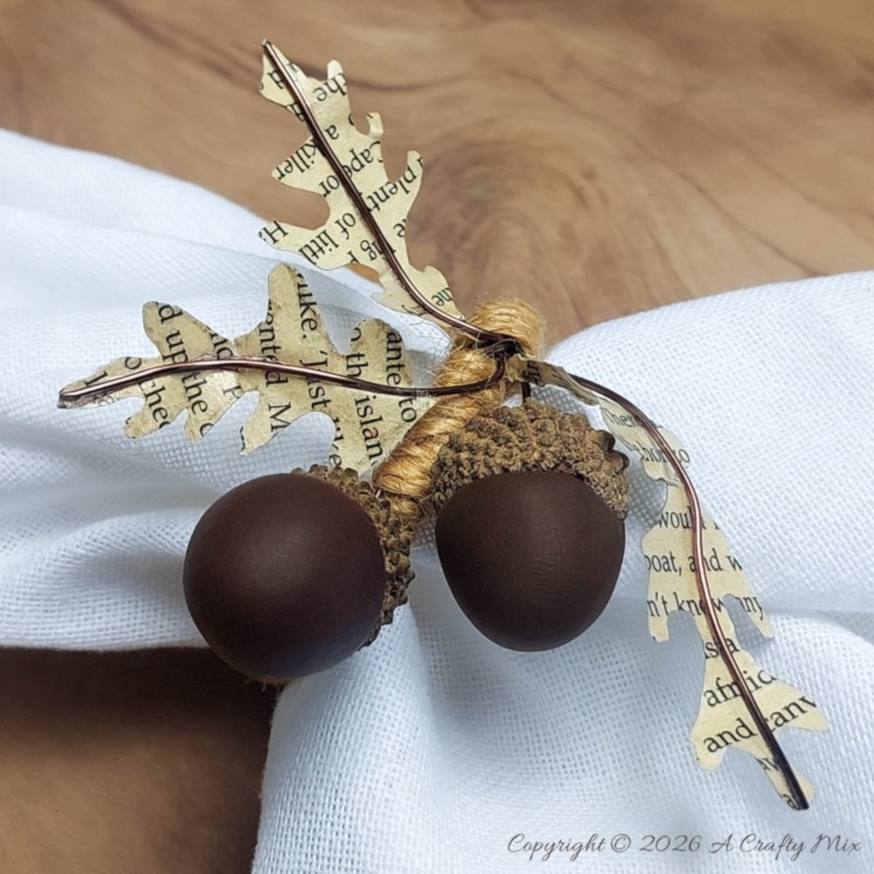 Handmade acorn napkin ring around a white cloth napkin with two brown clay acorns, real acorn caps, wired book-page leaves, and jute twine.