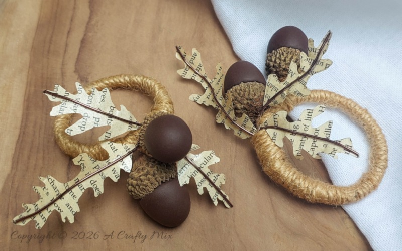 Two rustic DIY acorn napkin rings with clay acorns, real caps, book-page leaves, and twine on a wood surface beside a white napkin.