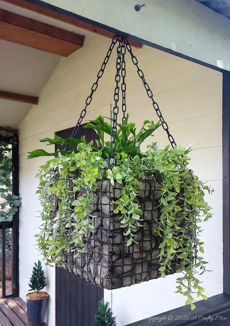 Large square gabion-style hanging planter with faux rocks and trailing plants hanging on a covered porch