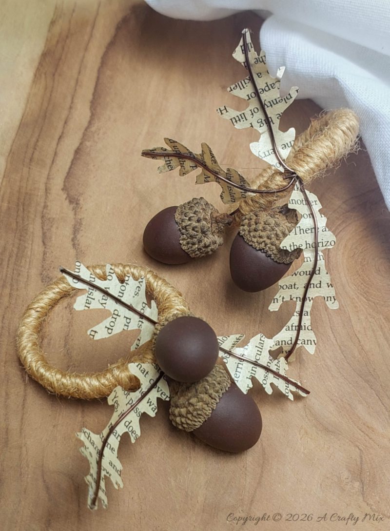 Close-up of two DIY acorn napkin rings with brown clay acorns, real acorn caps, copper wire, jute twine, and book-page leaves.