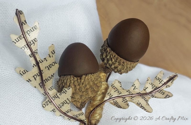 Close-up of book-page oak leaves wired with copper wire beside clay acorns, real acorn caps, and a twine-wrapped napkin ring.