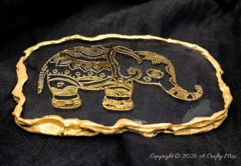 Gold elephant design embedded in a clear oval resin keepsake with an uneven textured gold border.